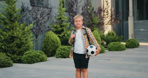 Portrait of confident blonde boy posing with footbal ball outdoors modern school Stock Footage 248615231