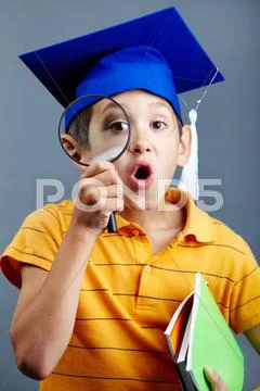 Portrait of curious boy in graduation hat looking through magnifying glass Stock Photos
