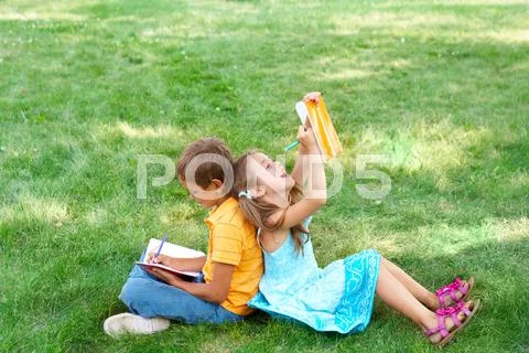 Portrait of cute kids seated on green grass with copybooks and pencils and drawi Stock Photos