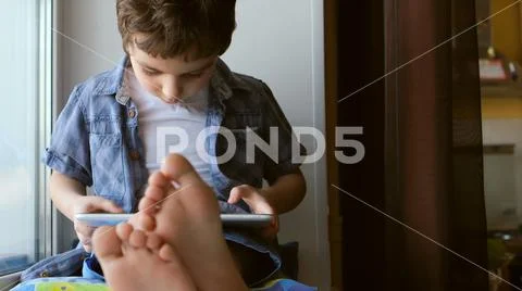 PORTRAIT: Cute little boy sits on a windowsill at home and touches a tablet PC Stock Photos