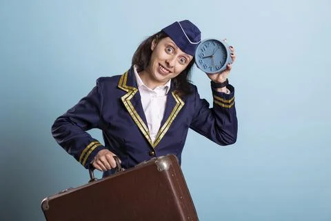 Portrait of female pilot holding suitcase and blue clock against studio backdrop 写真素材