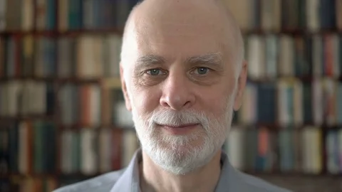 Portrait of good-looking handsome smiling senior man at home. Bookcase Stock Footage 74756624