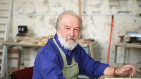 Portrait of a gray-haired carpenter at a table in the workshop with various Stock Photos