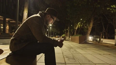 Portrait of a guy in white tshirt sitting with smartphone on a bench under city Stock Footage 155798985