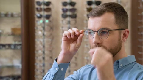 Portrait of handsome bearded guy picking new specs at optical shop, looking at Stock Footage 230924809