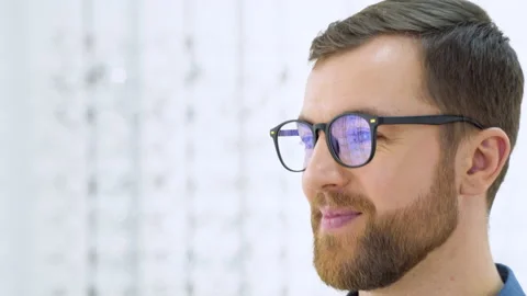 Portrait of handsome bearded guy picking new glasses at optical shop, looking at Stock Footage 231678127