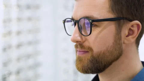 Portrait of handsome bearded guy picking new glasses at optical shop, looking at Stock Footage 231678581