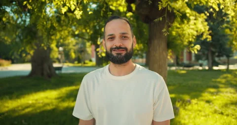 Portrait of a happy brunette guy with a beard in a white T-shirt in the park on Stock Footage 280578566