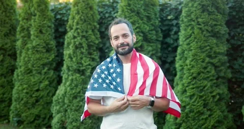 Portrait of a happy brunette guy with a beard wrapped in a US flag posing among Stock Footage 283832869