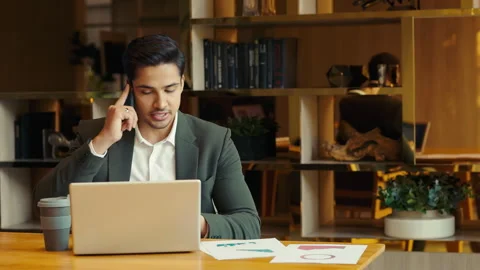Portrait of Indian manager with diagrams talks on cellphone at laptop indoors Stock Footage 305277953