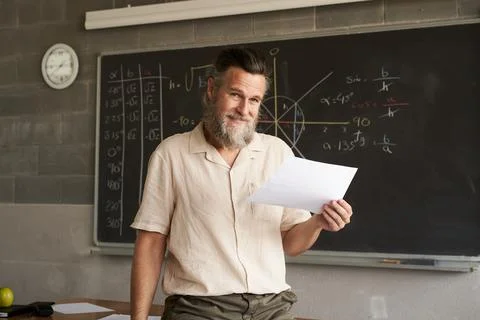 Portrait looking at camera of a male teacher in a classroom. In his hand he is Stock Photos