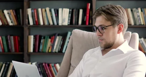 Portrait of man in glasses is working on computer on bookshelves background. Видео 131857687
