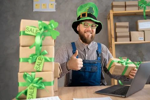 Portrait of man in leprecaun hat with boxes beside he uses a laptop to sell p Stock Photos