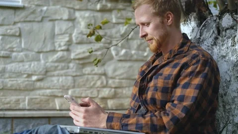 Portrait of a man with a smartphone sitting under a tree near the house Stock Footage 200825570