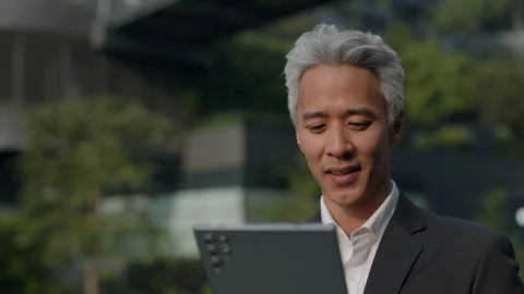 Portrait of mature Asian manager scrolls internet on tablet and stands in park Stock Footage 319892645