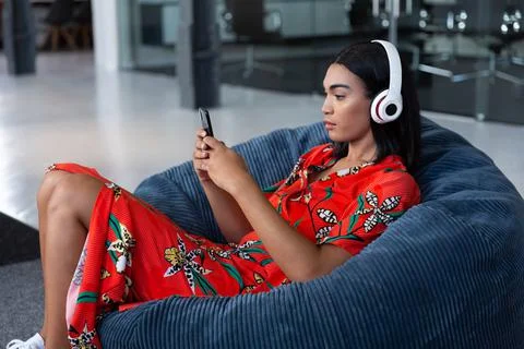 Portrait of mixed race businesswoman sitting in bean bag wearing headphones and Stock Photos