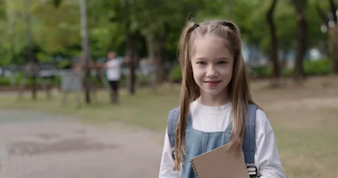 Portrait of primary class schoolgirl with notebook stands in park and smiles Stock Footage 346623436