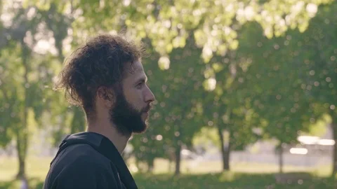 Portrait in a profile of a young bearded man standing in sun shining summer park Stock Footage 99493701