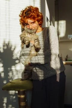 Portrait red-haired curly young woman with pet gray fluffy cat in arms, lit.. 스톡 사진