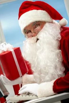 Portrait of santa claus holding gift in front of computer and looking at camera Stock Photos