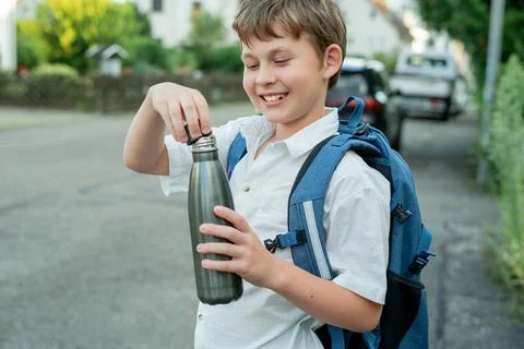 Portrait of a schoolboy with a backpack opens an iron bottle of water. Back t Stock Photos