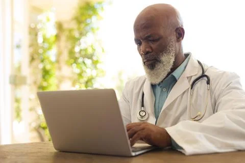 Portrait of senior african american male doctor in lab coat using laptop Stock Photos