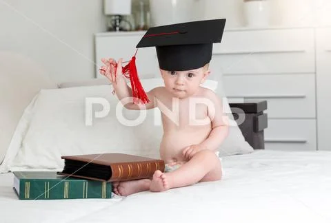 Portrait of smart baby boy in mortarboard cap sitting with books in bedroom Stock Photos