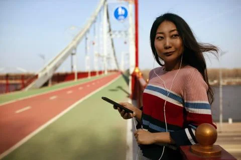 Portrait of a smiling asian fitness girl listening to music on bridge.