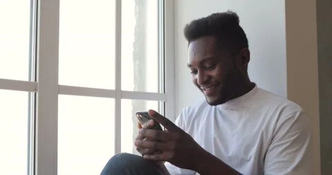 Portrait of a smiling black guy sitting near the windows, holding a smartphone Stock Footage 233323463