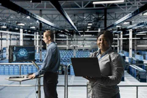 Portrait of smiling female engineer in server farm next to colleague using AI Foto stock