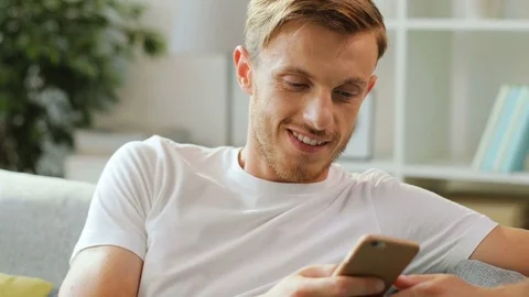 Portrait of smiling man in white t-shirt sitting on the sofa and sending sms Stock Footage 79997670