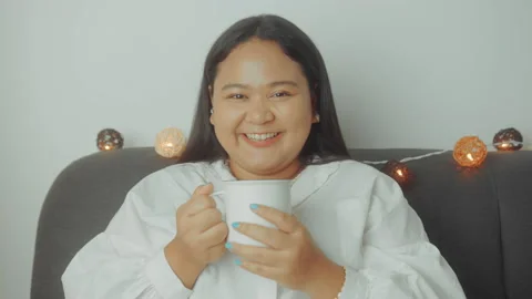 Portrait of a smiling plus size Asian woman sitting on a sofa and drink tea Stock Footage 153755358