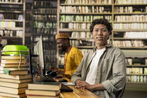 Portrait of smiling student sitting at library computer desk with stack of books Stock Photos