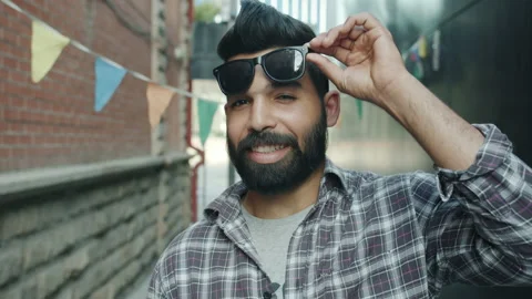 Portrait of stylish Arab guy raising sunglasses smiling and winking outdoors in Stock Footage 156760438