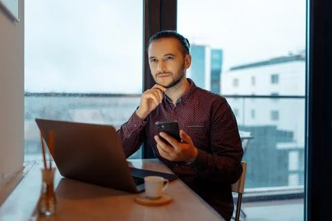 Portrait of thoughtful man working at laptop with smartphone in hand, backg.. Stock Photos