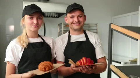 Portrait of two good looking young smiled male female bakers posing, croissants Stock Footage 144049055
