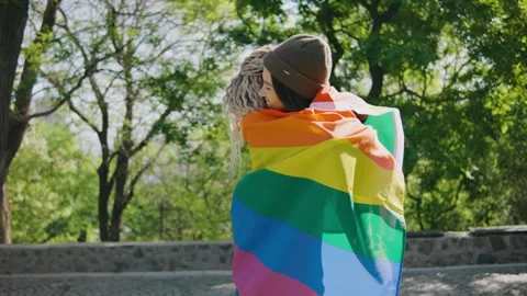Portrait of two smiling young lesbian women hugging in park under a rainbow flag Stock Footage 274320118