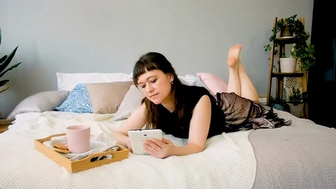 Portrait of a woman using a tablet and drinking coffee lying on a bed. 库存影片 127616337