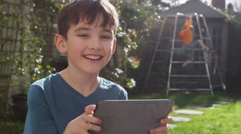 Portrait of a young boy playing on his touchscreen tablet in a garden on a brigh Stock Footage 47487383
