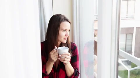 Portrait of young brooding girl sitting on the windowsill next to the window Stock Footage 146043304