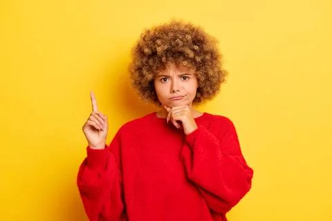 Portrait of young curly haired thinking woman, dressed in red jumper, makes Foto stock