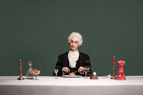 Portrait of young elegant man in peruke and vintage jacket sitting at table Stock Photos