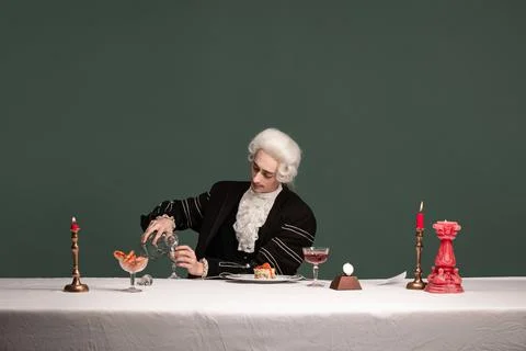 Portrait of young elegant man in peruke and vintage jacket sitting at table Stock Photos