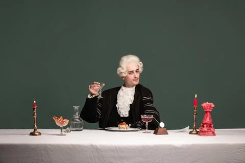 Portrait of young elegant man in peruke and vintage jacket sitting at table Stock Photos