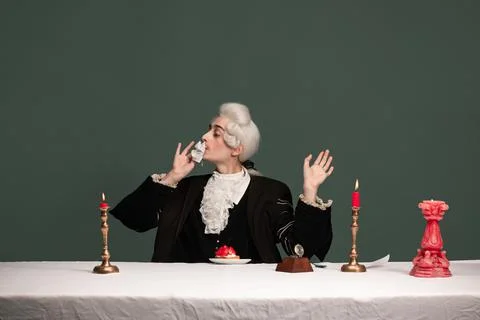 Portrait of young elegant man in peruke and vintage jacket sitting at table Stock Photos