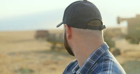 Portrait of the young farmer in a hat looking at combine working in the field Stock Footage 94449491
