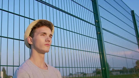 Portrait of a young guy in a hat against a metal grid, looking pensively toward Stock Footage 240660399