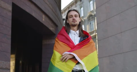 Portrait of young long haired man covered himself in rainbow flag while looking Stock Footage 136647152