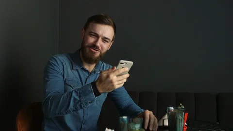 Portrait of a young man with a beard in a cafe. Blue shirt Man uses video call Vídeos de archivo 100599306