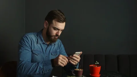 Portrait of a young man with a beard in a cafe. Blue shirt Man writes a message Vídeos de archivo 100601223